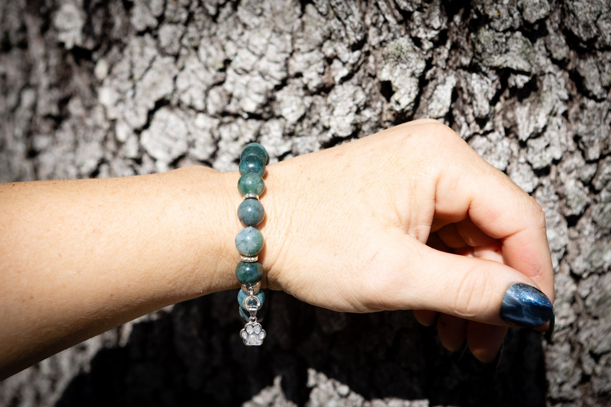 Hand wearing a green beaded bracelet with a small pawprint charm with crematin ashagainst a textured gray background